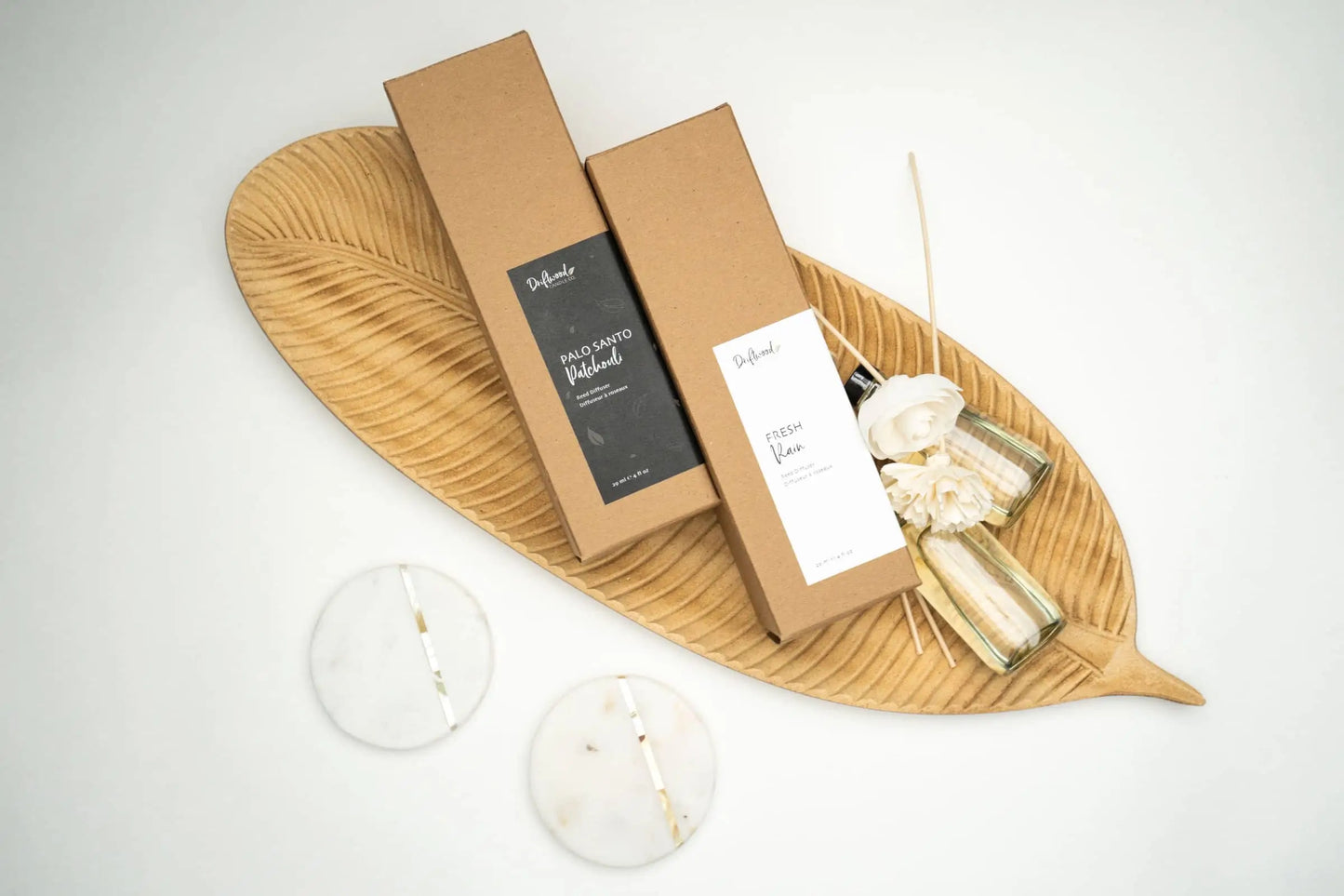 Overhead shot of two Driftwood Candle Co. Reed Diffuser sets on a decorative wooden leaf tray with sample bottles of fragrance oil and sola flowers next to them.