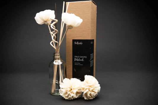 Sample Palo Santo Patchouli Reed Diffuser set on display. Set on a black backdrop.  Kraft box is labelled with the scent.  Next to the box is a sample of the reed diffuser set including two sola flowers, three reed stick and two curly reed sticks.  Between the sample set and the box are two other sola flowers sitting on their side.