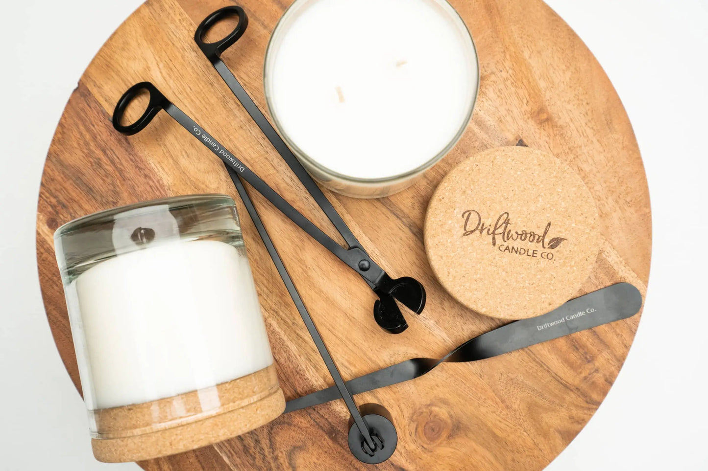 Decorative overhead shot of two Driftwood Candle Co candles with cork lids on a round table riser shelf with a wick trimmer, snuffer and candle dipper 